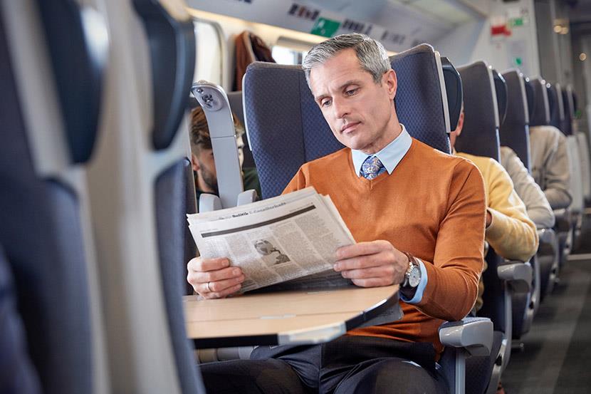 man in orange sweater sitting on a train reading a papers